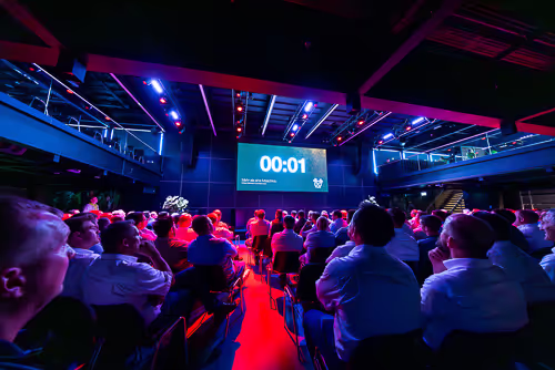 Publiek zit in een zaal op een bood tijdens een evenement met een groot scherm waarop een timer staat met 00:01.